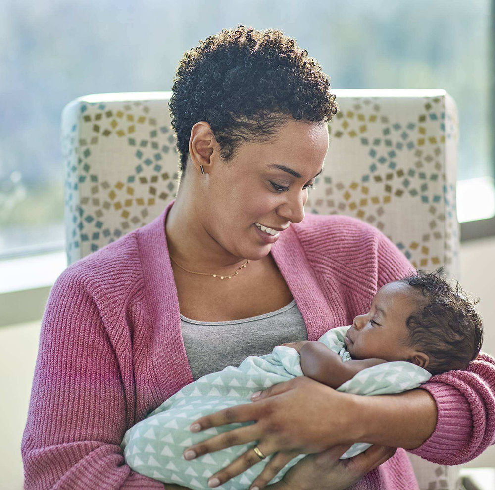 A mother smiling warmly while cradling her sleeping swaddled newborn baby