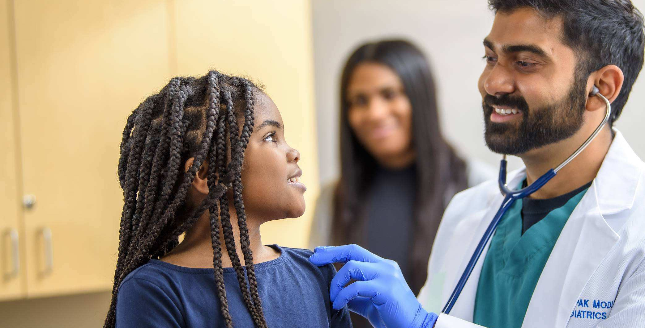 A male pediatrician with a stethoscope examining a smiling young girl.