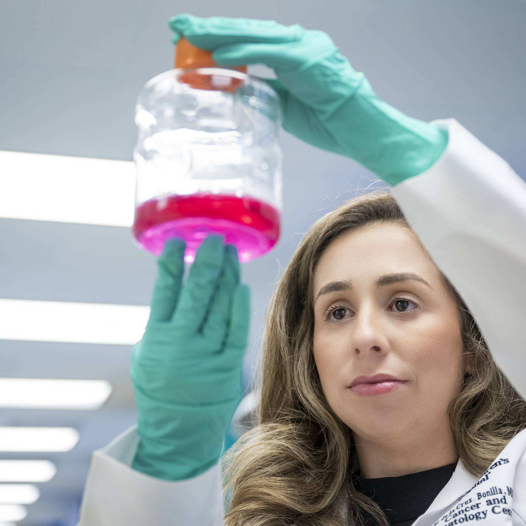 A female scientist in a lab coat wearing teal gloves holds up a glass container filled with bright pink liquid to examine it