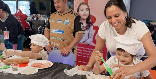 Two young children wearing white chef hats making food at a table with help from adults during a hospital event
