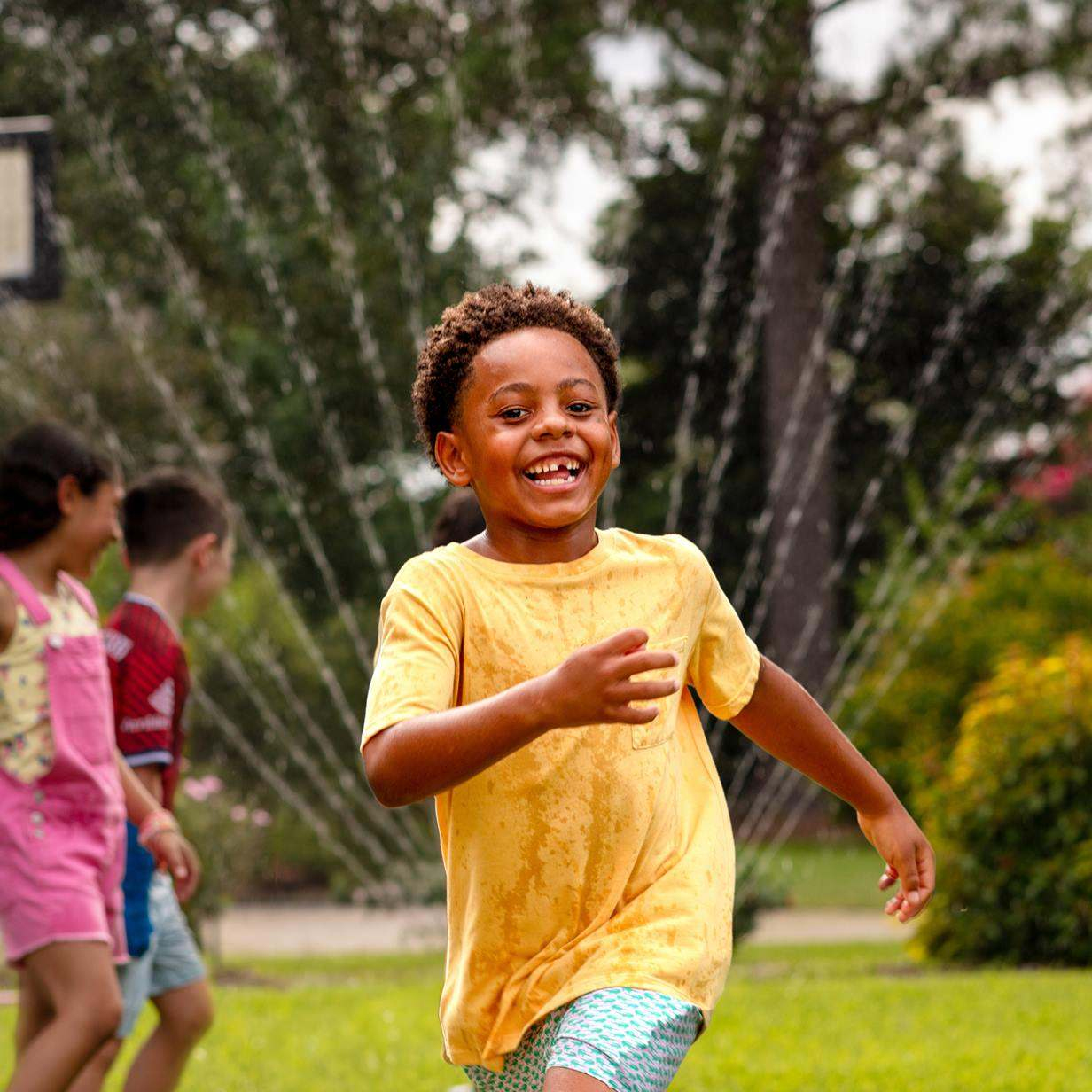 A smiling young boy in a yellow shirt running across a grassy lawn with water spraying from a sprinkler behind him