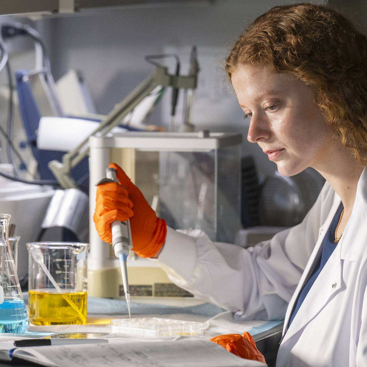 A red-haired scientist wearing orange gloves carefully using a pipette to transfer liquid into a multi-well plate