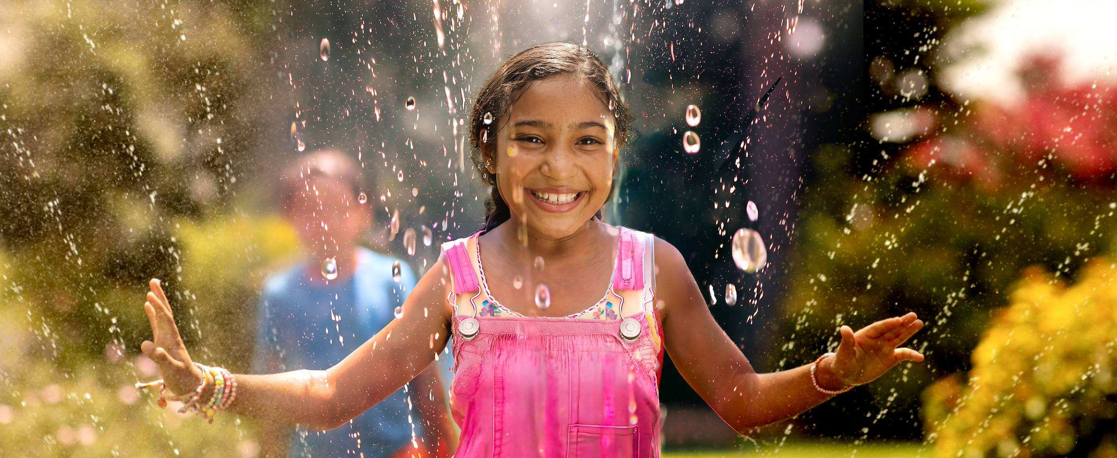 A smiling young girl in overalls playing outdoors under water splashing from a sprinkler.