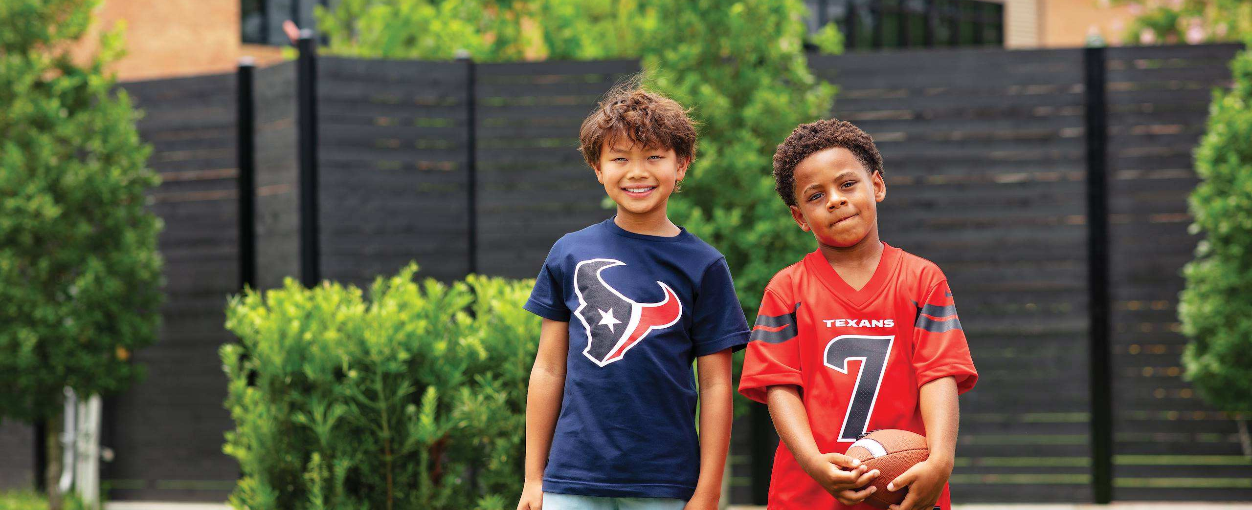 Two young boys wearing Houston Texans sports gear smiling together outdoors while holding a football.