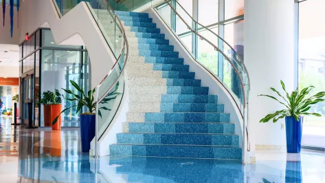 The inside of the West Austin building, showing a modern indoor staircase with blue gradient steps and glass railings, surrounded by large windows, vibrant green plants in blue and orange pots, and a glossy, reflective floor.