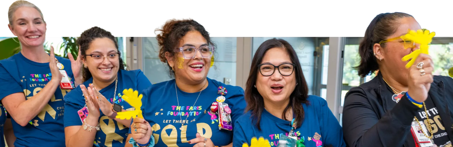 Five women stand together smiling and clapping while holding yellow paper flowers. They wear blue t-shirts that say "Let There Be GOLD" and appear to be at a cheerful event or celebration.