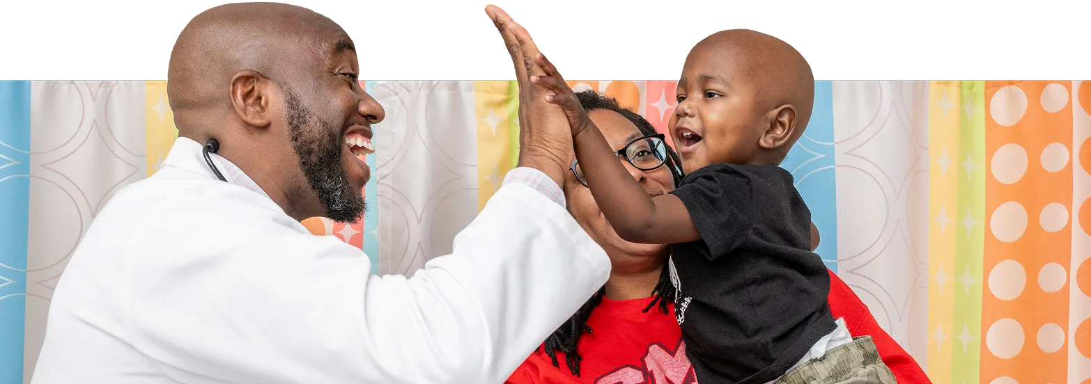 A smiling doctor gives a high-five to a happy toddler held by an adult woman, all in front of colorful patterned curtains.
