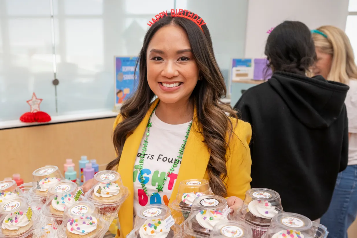 A smiling woman wearing a "Happy Birthday" headband and yellow blazer holds cupcakes at a party. She stands in front of a table with more cupcakes, with people and decorations in the background.