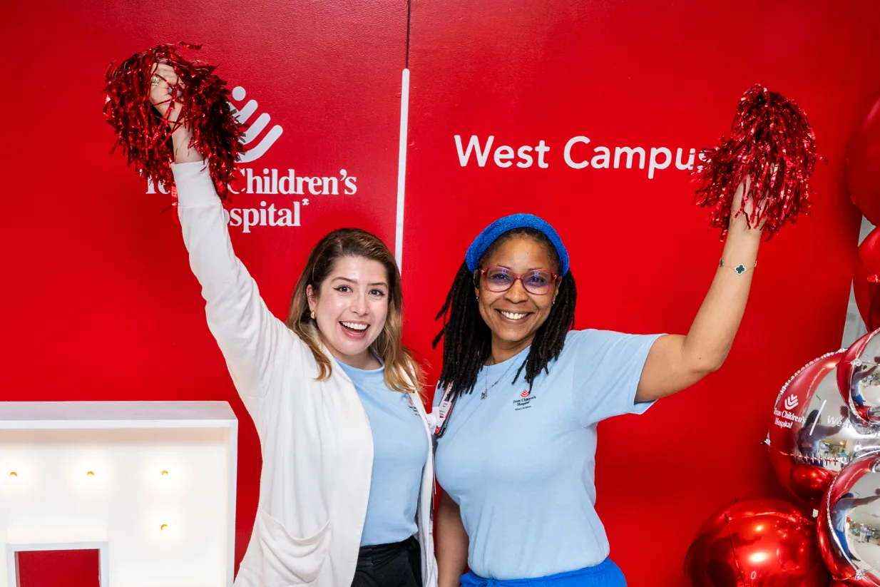 Two women smiling and holding red pom-poms stand in front of a bright red wall with the Texas Children’s Hospital West Campus logo. Both wear light blue shirts, and silver balloons are visible on the right.