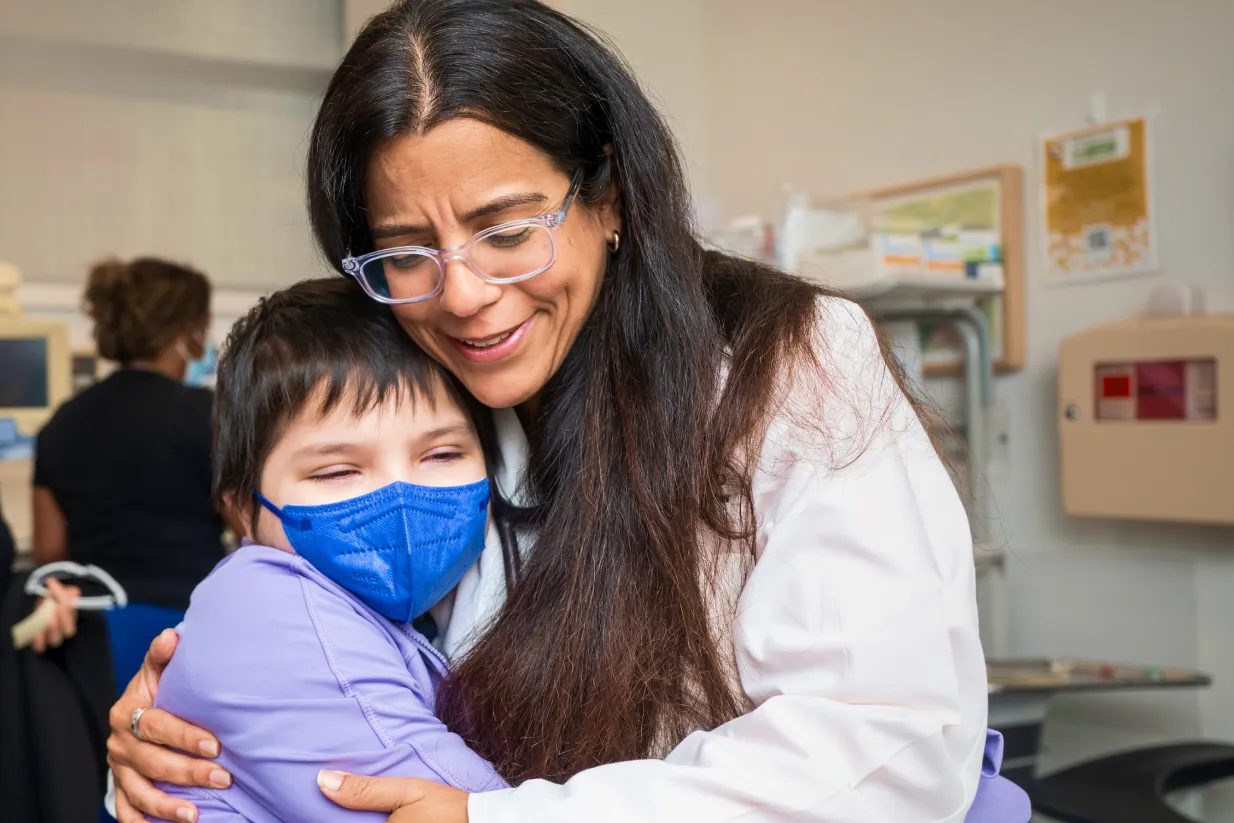 A woman in a white coat hugs a smiling child wearing a blue mask and purple shirt in a medical setting. The woman appears happy, and the child looks content, with medical equipment visible in the background.