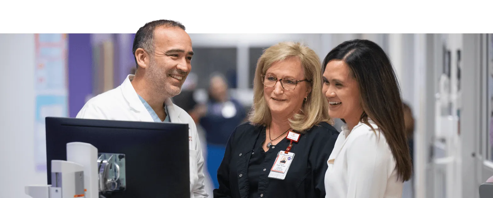 Three healthcare professionals, two women and one man, stand together smiling and looking at a computer monitor in a bright medical facility hallway.