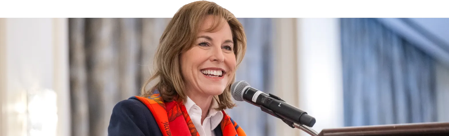 A woman with shoulder-length brown hair smiles while speaking into a microphone. She is wearing a dark blazer and a red patterned scarf, standing indoors with blurred curtains in the background.