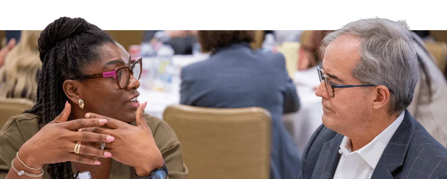A woman and a man sit at a table in conversation during a professional event. The woman gestures with her hands while the man listens attentively. Other people are visible in the blurred background.