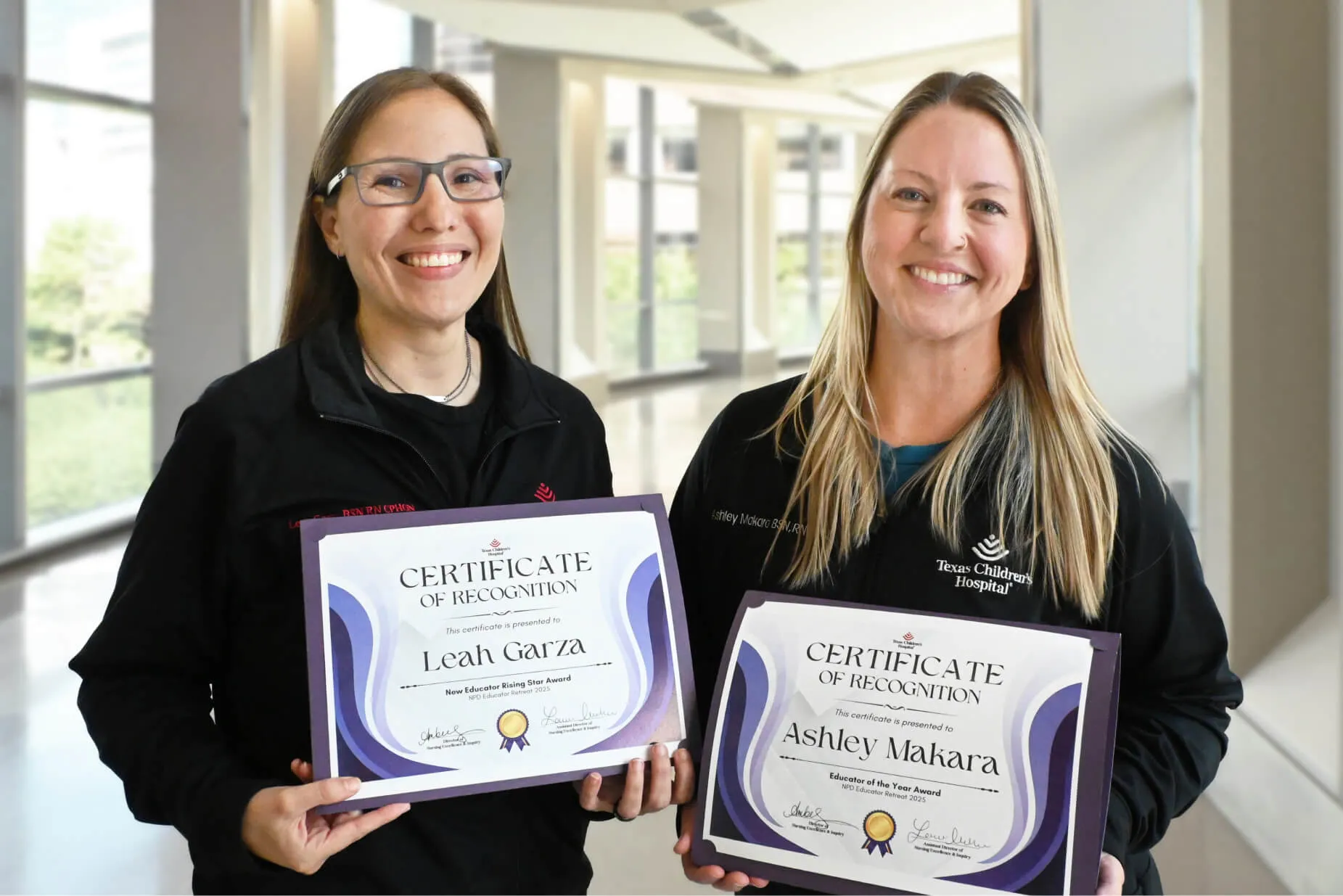 Two women are smiling and holding certificates of recognition in a bright, modern hallway. The certificates display the names Leah Garza and Ashley Makara. Both women wear black jackets and appear happy.