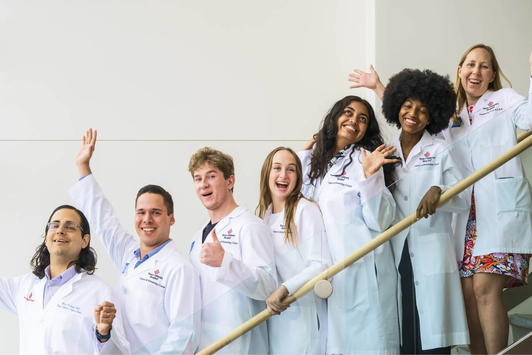 Seven medical professionals in white lab coats stand on a staircase, smiling and waving enthusiastically. The group is diverse in gender and ethnicity, and they appear happy and celebratory in a bright, modern space.