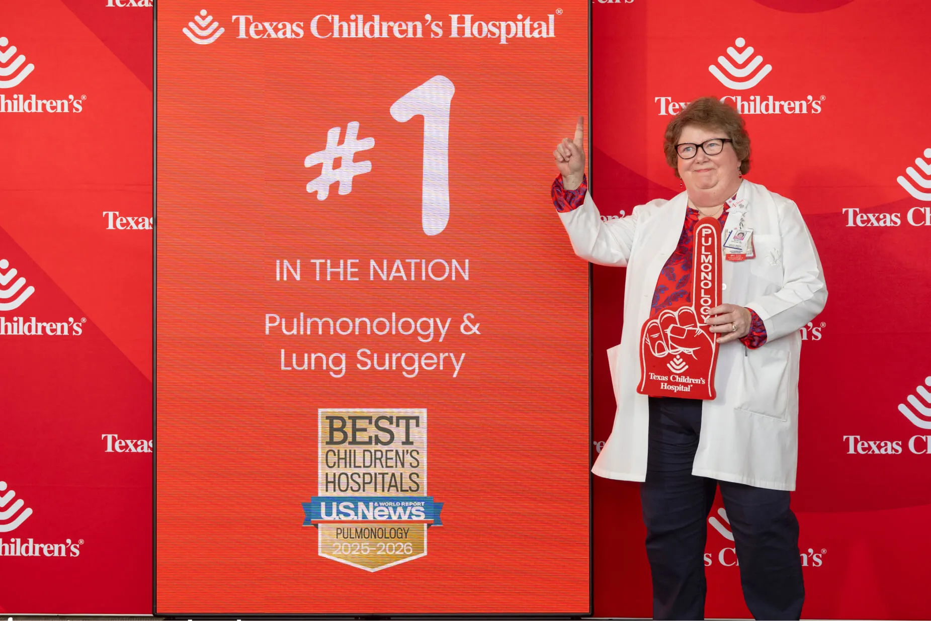 A doctor in a white coat stands beside a sign that reads “#1 in the nation Pulmonology &amp;amp; Lung Surgery” at Texas Children’s Hospital, holding a foam finger and smiling. The background is red with hospital logos.
