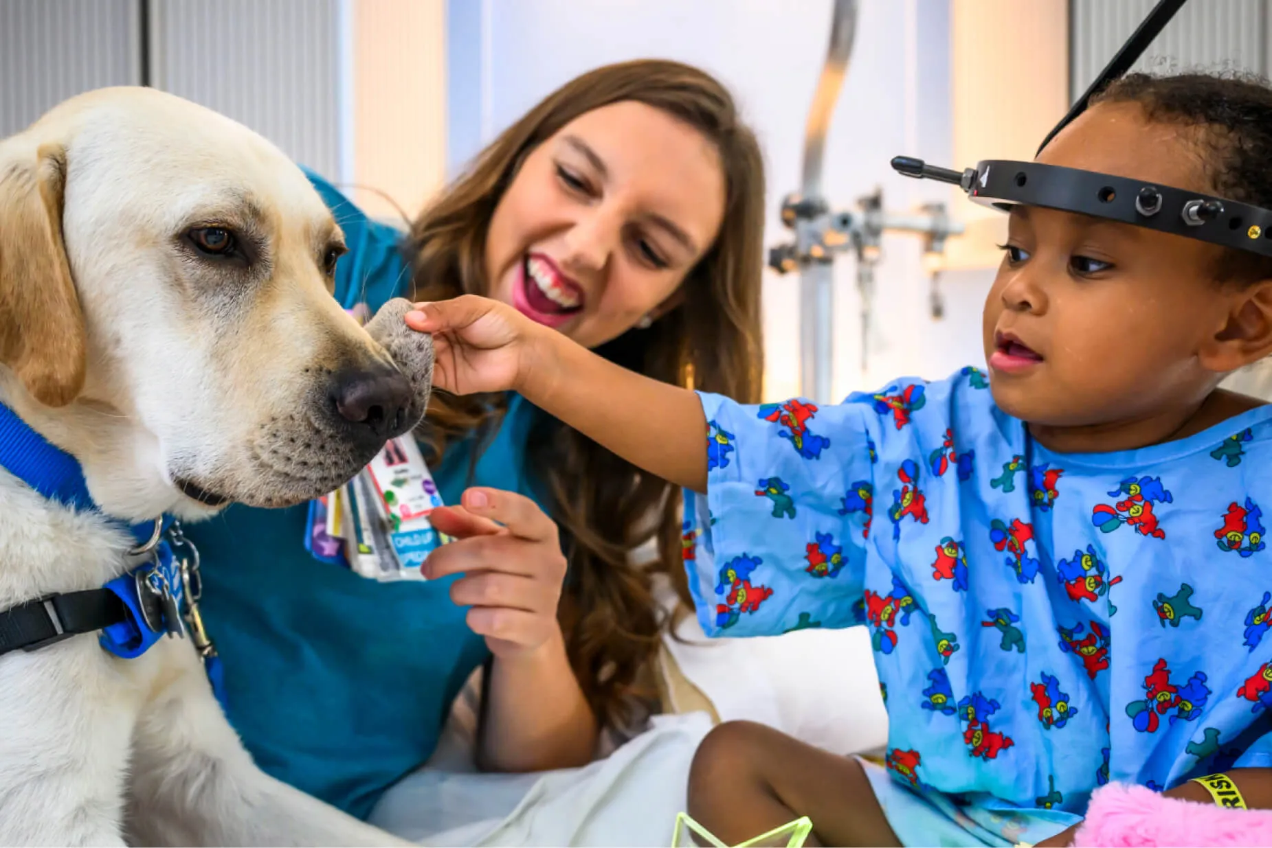 A child in a hospital gown pets a Labrador dog while a woman, possibly a nurse or caretaker, smiles encouragingly beside them in a hospital room.