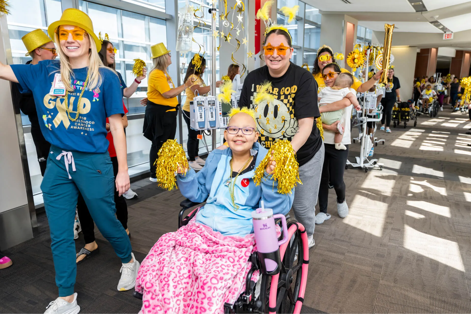 A smiling child in a wheelchair, wearing a pink blanket, is surrounded by people in cheerful yellow hats and pom-poms, parading indoors. Medical staff and other children join the lively, supportive event.