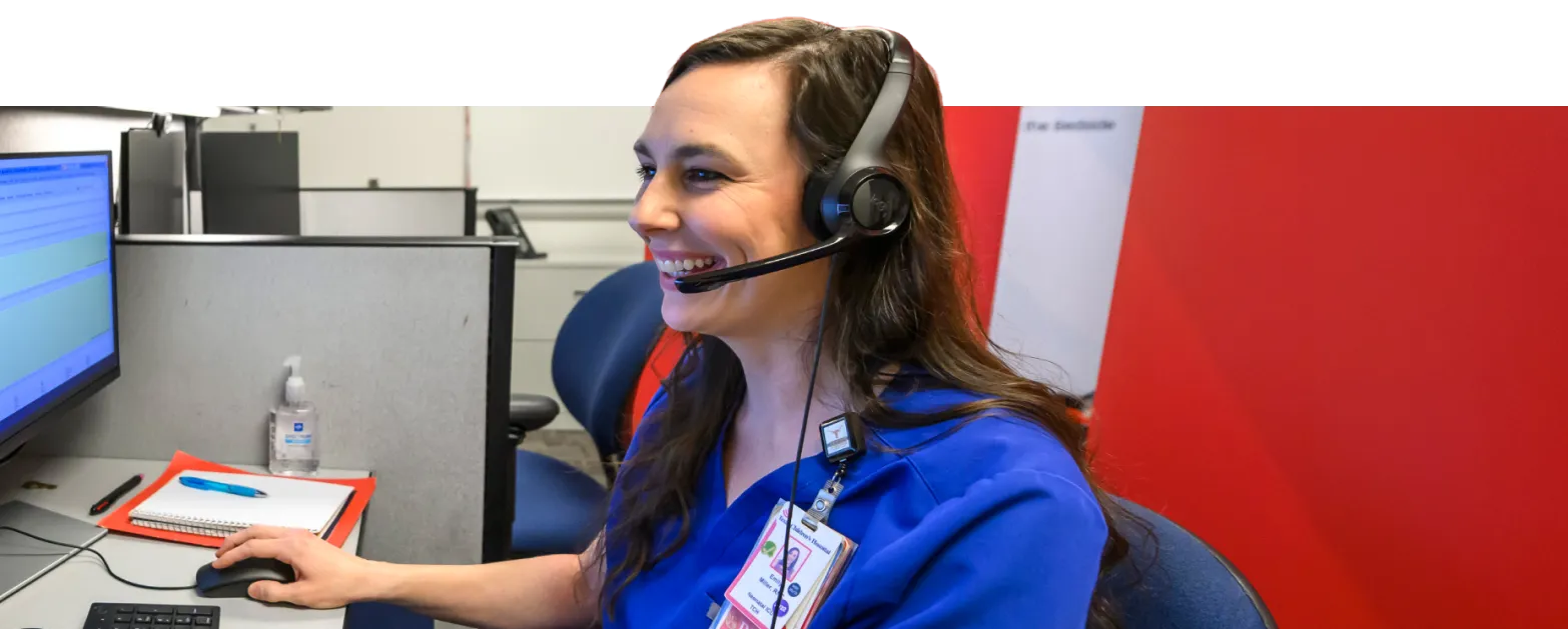 A woman wearing a blue uniform and headset smiles while working at a computer in an office cubicle. A notepad, pen, and hand sanitizer are visible on her desk.