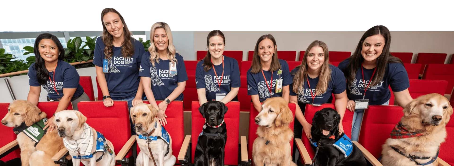 Four women in red shirts kneel on grass, each with a golden retriever wearing a red hat and matching red, white, and blue plaid outfits. All are smiling, posing together outdoors.