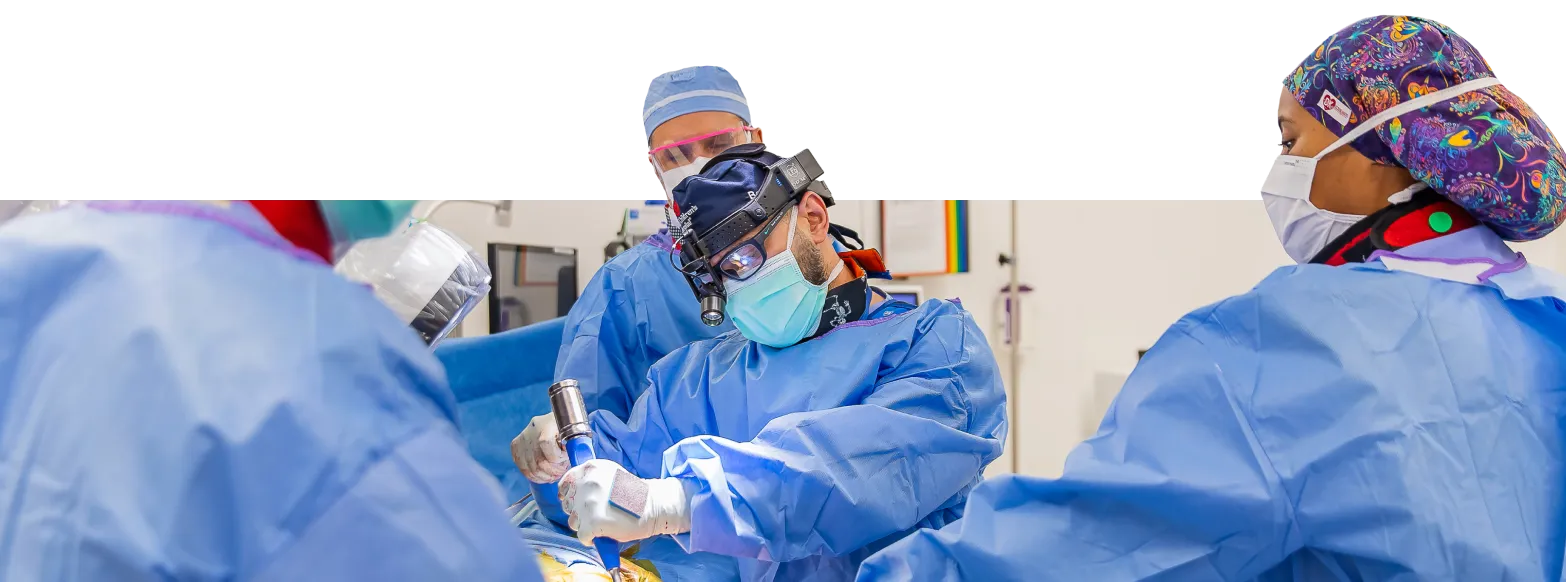 Three healthcare professionals wearing surgical masks, gowns, and caps focus on a procedure in a hospital setting. Medical equipment and monitors are visible in the background.