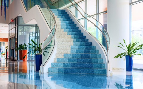 The inside of the West Austin building, showing a modern indoor staircase with blue gradient steps and glass railings, surrounded by large windows, vibrant green plants in blue and orange pots, and a glossy, reflective floor.